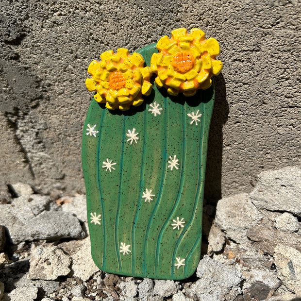 Flat ceramic cactus sculpture, green with white star burst meant to emulate spikes. At the top are 2 bright yellow blossomed flowers.