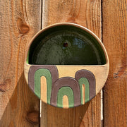 Ceramic circular vessel, terracotta color with brown and green curved lines, abstract and geometric. Interior of vessel is olive green and hangs on a wall.
