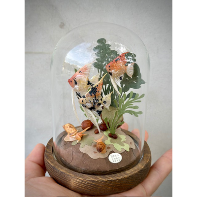 Mixed media sculpture diorama in a glass cloche. A school of orange and black speckled angel fish swim around cut paper leaves. Snails sit on the bottom of the scene.