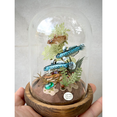 Mixed media sculpture of 2 bright blue fish, made out of clay and one brown fish, swimming around paper cut leaves. Scene is encased in a glass cloche.