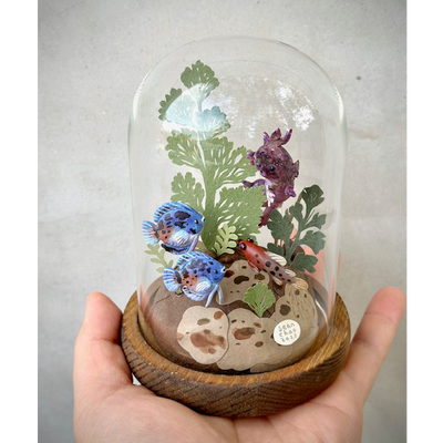 Mixed media sculpture of various colorful fish, made out of clay and swimming around cut paper leaves and plants. Fish are blue, red and purple. Scene is encased in a glass cloche.