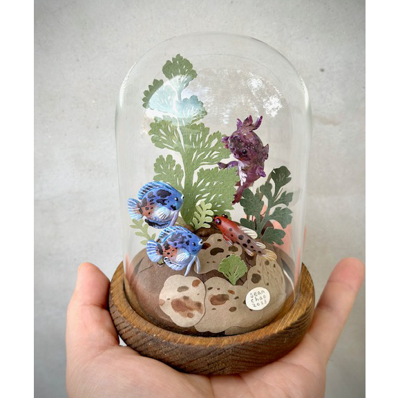 Mixed media sculpture of various colorful fish, made out of clay and swimming around cut paper leaves and plants. Fish are blue, red and purple. Scene is encased in a glass cloche.