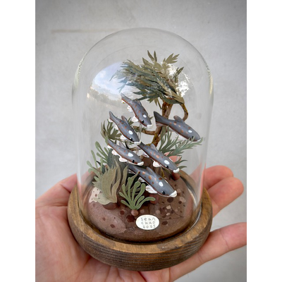 Sculpture of a school of grey fish, made from clay, swimming side by side one another. Cut paper sea grass and greenery is around them. The scene is inside of a glass cloche.