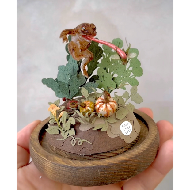 Mixed media diorama of a frog, jumping up and extending a large tongue to catch a fly. It's above a small patch of pumpkin and other ground vegetables.