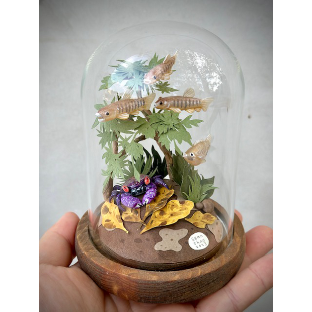 Mixed media sculpture diorama in a glass cloche. A school of brown fish made of clay swim around cut paper plants. At the bottom of the scene is a purple crab.