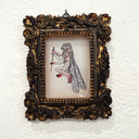 An ornate, vintage-style baroque frame with gold and dark brown detailing, containing a small illustration of a winged fairy holding a dagger, mounted on a textured white wall.