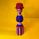 Colorful wooden sculpture that resembles a candle holder in shape (thin and tall) with the face of a woman wearing striped clothing.