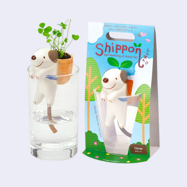 Ceramic white dog with brown eyes and cartoon smile, positioned over a glass of water with its arms hanging on the lip of the glass. On its back is a ceramic pack which grows clover. It stands next to its product packaging.