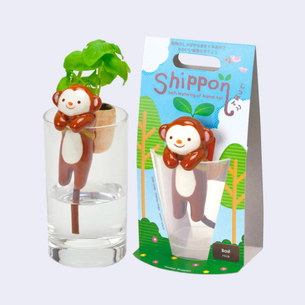 Ceramic brown monkey with a smiling cartoon face, positioned over a glass of water, its arms hanging onto the lip. On its back is a ceramic bag with basil growing out of it. Positioned next to its product packaging.