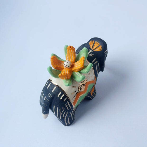 Matte surfaced ceramic sculpture of a black elephant with long white and gold tusks. Its body is patterned with cream colored lines, orange poppy flowers and It wears a fabric designed like mountains with a setting gold sun. Atop its back is an orange flower with green leaves.