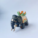 Matte surfaced ceramic sculpture of a black elephant with long white and gold tusks. Its body is patterned with cream colored lines, orange poppy flowers and It wears a fabric designed like mountains with a setting gold sun. Atop its back is an orange flower with green leaves.