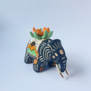Matte surfaced ceramic sculpture of a black elephant with long white and gold tusks. Its body is patterned with cream colored lines, orange poppy flowers and It wears a fabric designed like mountains with a setting gold sun. Atop its back is an orange flower with green leaves.