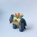 Matte surfaced ceramic sculpture of a black elephant with long white and gold tusks. Its body is patterned with cream colored lines, orange poppy flowers and It wears a fabric designed like mountains with a setting gold sun. Atop its back is an orange flower with green leaves.