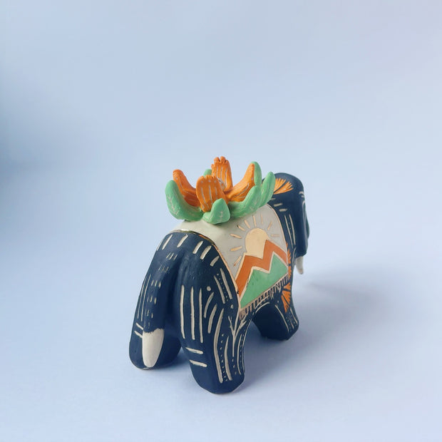 Matte surfaced ceramic sculpture of a black elephant with long white and gold tusks. Its body is patterned with cream colored lines, orange poppy flowers and It wears a fabric designed like mountains with a setting gold sun. Atop its back is an orange flower with green leaves.