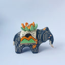 Matte surfaced ceramic sculpture of a black elephant with long white and gold tusks. Its body is patterned with cream colored lines, orange poppy flowers and It wears a fabric designed like mountains with a setting gold sun. Atop its back is an orange flower with green leaves.