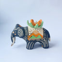 Matte surfaced ceramic sculpture of a black elephant with long white and gold tusks. Its body is patterned with cream colored lines, orange poppy flowers and It wears a fabric designed like mountains with a setting gold sun. Atop its back is an orange flower with green leaves.