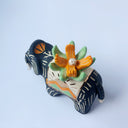 Matte surfaced ceramic sculpture of a black elephant with long white and gold tusks. Its body is patterned with cream colored lines, orange poppy flowers and It wears a fabric designed like mountains with a setting gold sun. Atop its back is an orange flower with green leaves.