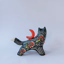 Ceramic figurine of a black wolf with white line detailing and red floral patterning. it holds a red tassel in its mouth and has a red crescent moon on its back.