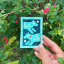 A hand holds an artwork featuring two stylized teal foxes on a dark teal background with white decorative border details, set against a lush green garden with pink flowers.