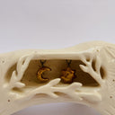 A cream colored ceramic wolf, with an opening in its body that holds gold charms, hanging from wire. Surrounded the opening are ceramic vine shapes.