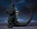 Godzilla action figure posed standing on a dark textured surface against a dramatic cloudy blue-gray background. The figure is dark gray with white dorsal spines and small gold/yellow claw tips.