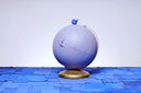 A globe, painted entirely light purple with a closed eye, calm facial expression. A small blue wooden snail rests atop the globe, which sits on a gold base.