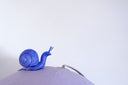 A globe, painted entirely light purple with a closed eye, calm facial expression. A small blue wooden snail rests atop the globe, which sits on a gold base.