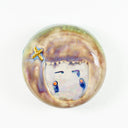 Round ceramic tile of a head - with small eyes and round cheeks. Around their head is a cropped haircut with an "x" hairbow.