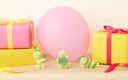 Three small figurines with birthday hats and a pink balloon on a wooden surface.
