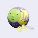 Small keychain of a Smiski, a simplistic round headed green character with a small face. It attempts to do a backwards arch, its positioned next to its capsule packaging.