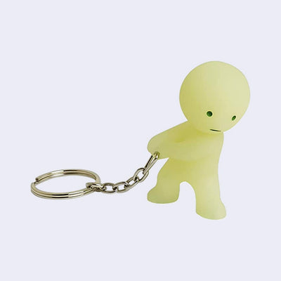 Smiski, a round headed character with simple features and a plain, serious look. Its positioned as if it's carrying something heavy, with a keychain attached to its hands.