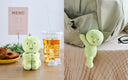 Green plush toy on a table with a drink and food, and another plush toy hanging from a bag.