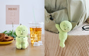 Green plush toy on a table with a drink and food, and another plush toy hanging from a bag.