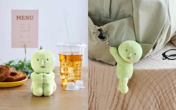 Green plush toy on a table with a drink and food, and another plush toy hanging from a bag.