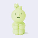 Sitting Smiski, a small plastic character, light green with a circular head and human like body. It has its knees pulled to its chest and a cat lays atop its head.