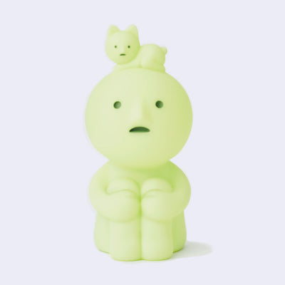 Sitting Smiski, a small plastic character, light green with a circular head and human like body. It has its knees pulled to its chest and a cat lays atop its head.