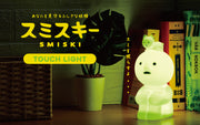 Sitting Smiski, a small plastic character, light green with a circular head and human like body. It has its knees pulled to its chest and a cat lays atop its head. Whole sculpture is a light, emitting a soft greenish glow.