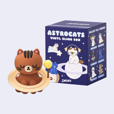 Vinyl figure of a brown cartoon cat, sitting with its eyes closed and rings around it like it is a planet. It sits in front of its product packaging.