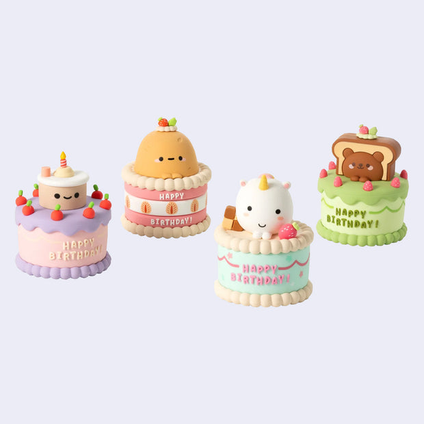 Set of four small, cute birthday cake figurines with cartoon faces on a light purple background.