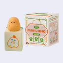 Small figurine of a chunky mahjong tile with a cartoon potato character sitting atop of it, in front of its product packaging.