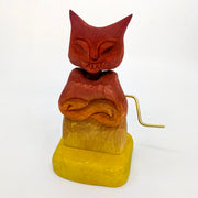 Whittled wooden sculpture of a red and yellow gradient devil with a cat like face and crossed arms. A gold crank comes out of the side.