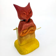 Whittled wooden sculpture of a red and yellow gradient devil with a cat like face and crossed arms. A gold crank comes out of the side.