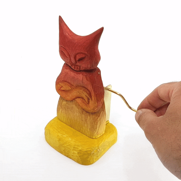 Gif of a red and yellow gradient wooden devil with a cat like face and crossed arms. Its head nods up and down, in sync with a gold crank being turned.