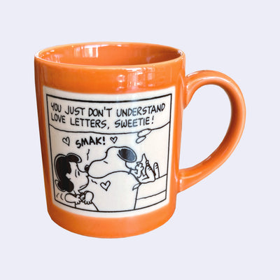 Orange mug with a cartoon design featuring Snoopy and a Lucy, with Snoopy giving Lucy a kiss on the nose.