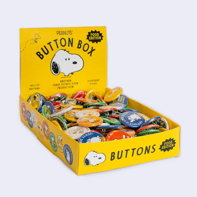 Yellow box filled with assorted tin buttons with graphics of food themed Snoopy illustrations on them. 
