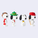 Four small Snoopy figurines in different outfits, arranged side by side on a plain light gray background. One wears a brown aviator hat and red scarf, one wears a green ranger hat with a yellow band, one wears a black-and-white outfit with a yellow bow tie, and one wears a red cap and holds a wooden bat.