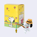 Snoopy figurine with a hat next to a yellow Snoopy-themed box on a white background
