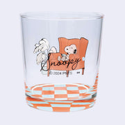 Clear glass with Snoopy design sitting on an orange chair next to a plant atop of a checkered floor. "Snoopy" is written in cursive on the back.