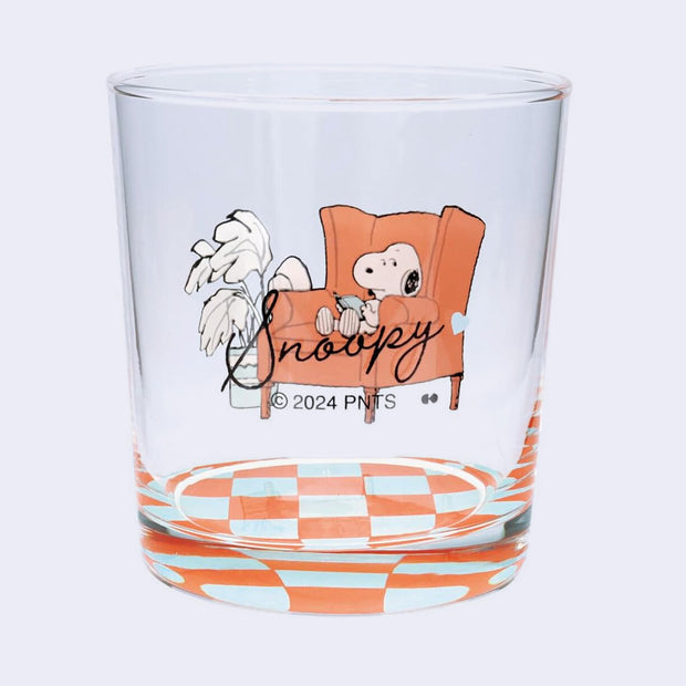 Clear glass with Snoopy design sitting on an orange chair next to a plant atop of a checkered floor. "Snoopy" is written in cursive on the back.