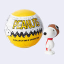 A Peanuts collectible character surprise ball next to a small figurine of Snoopy wearing a brown aviator hat and red scarf, on a light gray background.
